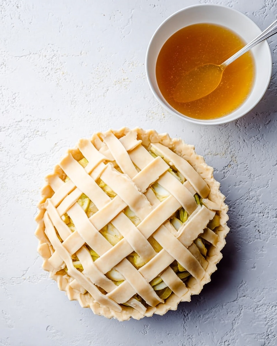 The image shows a round pie with a lattice crust on top made of wide, pale dough strips woven in a crisscross pattern. The edges of the pie crust are pinched and crimped, giving a decorative border. The filling underneath the lattice is slightly visible and looks light yellow. Next to the pie, there is a white bowl filled with a golden brown liquid and a spoon resting inside it. The pie and bowl are placed on a white marbled surface, creating a clean and simple background. photo taken with an iphone --ar 4:5 --v 7