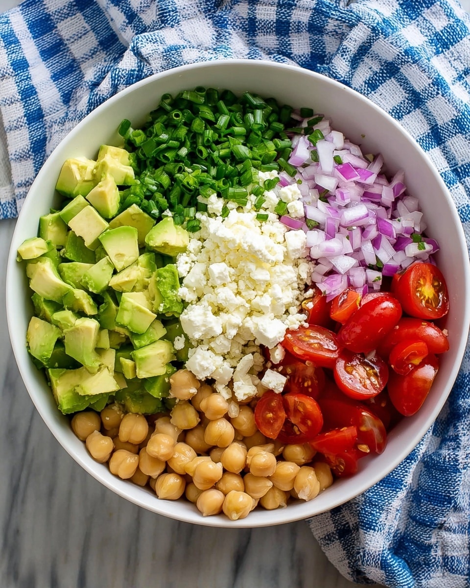 A white bowl holds five separate layers of food arranged side by side. Starting from the left, there are bright green avocado cubes with smooth texture, next to a pile of finely chopped dark green chives. At the center top, there is a soft, crumbly white cheese layer. To the right of the cheese, finely diced red onions add a purple hue, and on the far right, glossy red cherry tomato halves with juicy insides complete the arrangement. At the bottom right, there is a layer of round, light tan chickpeas with a smooth surface. The bowl is resting on a white marbled texture with a blue and white checkered cloth partly visible behind it. Photo taken with an iphone --ar 4:5 --v 7