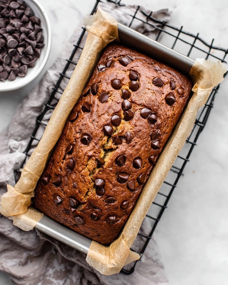 A rectangular chocolate chip banana bread loaf in a silver baking pan lined with light brown parchment paper on both sides. The top of the loaf is golden brown with a slightly cracked surface, studded with many shiny, melted dark chocolate chips spread evenly. The loaf sits on a black wire cooling rack placed over a white marbled surface. In the background, there is a white bowl filled with dark chocolate chips on a light gray cloth. Photo taken with an iphone --ar 4:5 --v 7