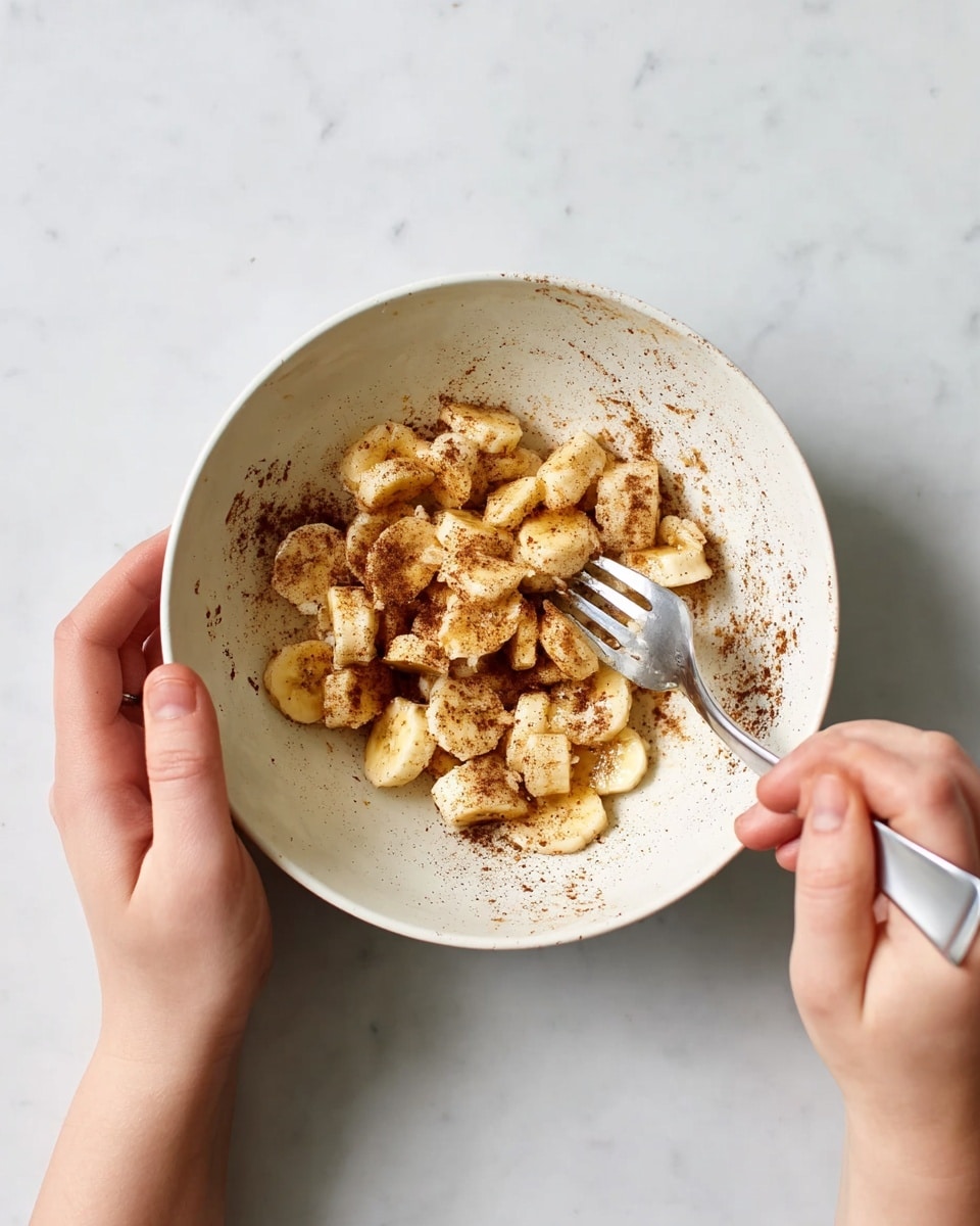 A woman’s hand holds a white bowl filled with small pieces of crushed banana, lightly coated with brown cinnamon powder, while another woman’s hand uses a fork to mash the banana. The banana pieces have a soft, slightly mushy texture with some smooth parts, and the cinnamon is unevenly spread inside the bowl. The bowl sits on a white marbled surface. photo taken with an iphone --ar 4:5 --v 7