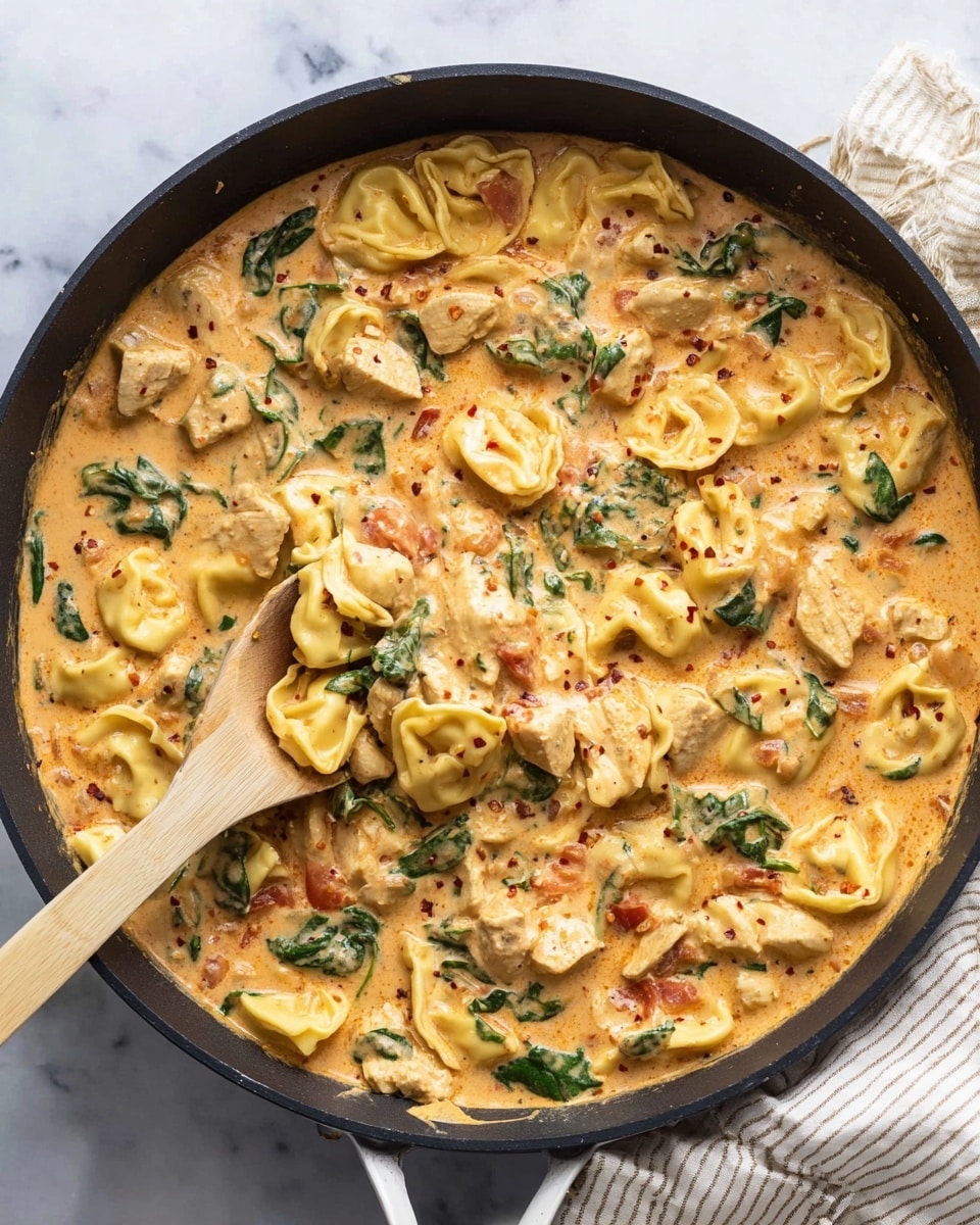 A black pan filled with creamy orange sauce holding three main layers: light golden tortellini pasta shapes with ruffled edges, tender white pieces of cooked chicken scattered evenly, and dark green spinach leaves sliced roughly over the top. Mixed in the sauce are visible red specks of seasoning, giving texture to the creamy surface. A wooden spatula with a white handle rests inside the pan, partially lifting some pasta and chicken. The background is a soft white marbled surface with a light-colored striped cloth near the pan's handle. Photo taken with an iphone --ar 4:5 --v 7