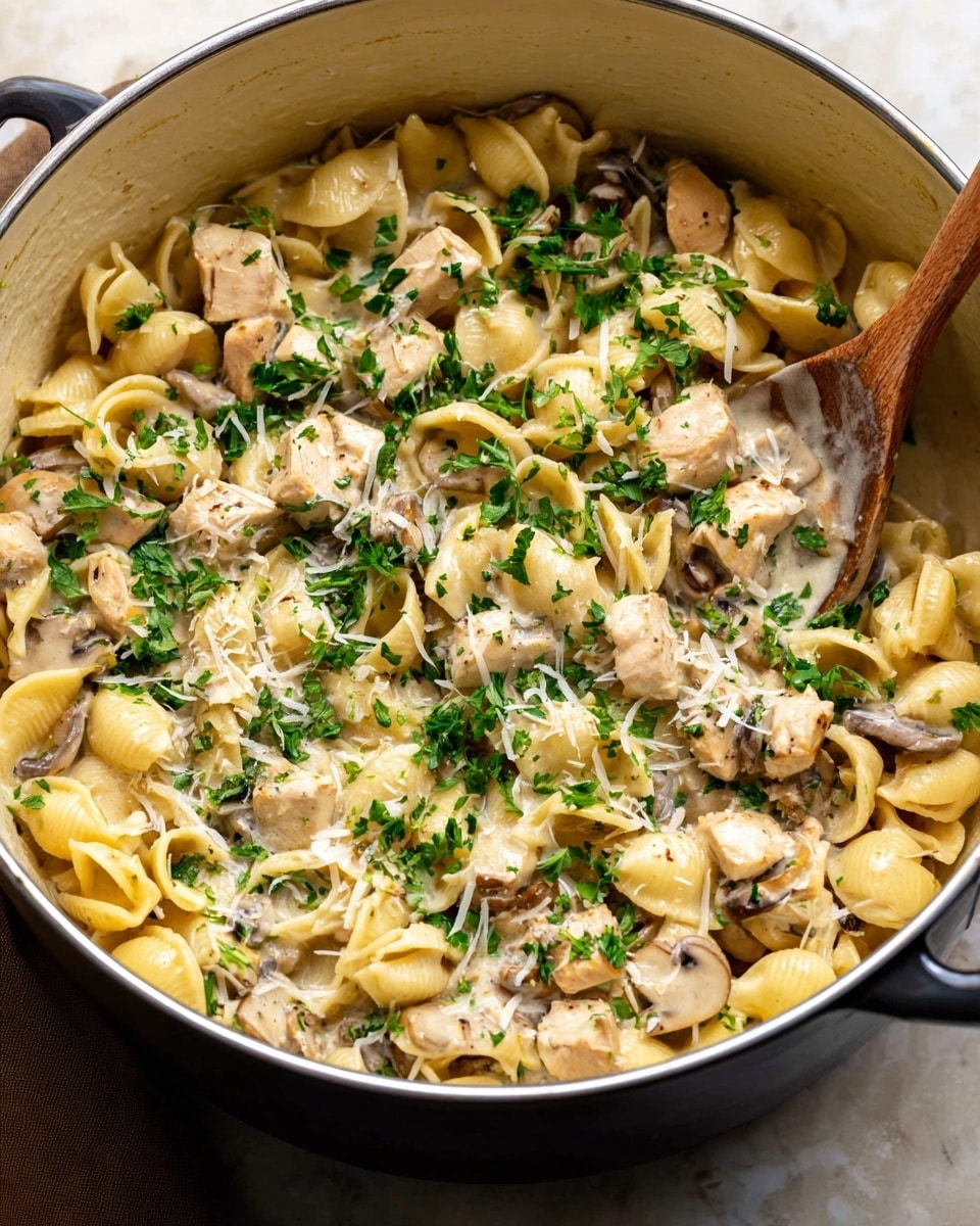 A large pot filled with creamy pasta made with small shell-shaped noodles mixed with light beige chicken pieces and brown mushroom slices. The pasta is coated in a smooth cream sauce with visible herbs. The top layer is sprinkled with finely chopped bright green fresh parsley and small shreds of pale grated cheese. The pot sits on a white marbled surface with a wooden spoon partially stirring the food inside. photo taken with an iphone --ar 4:5 --v 7