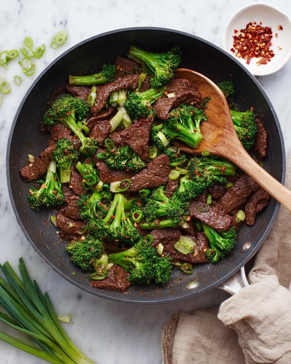 The image shows a black frying pan filled with cooked beef strips and bright green broccoli florets mixed together. The beef strips are dark brown with a slightly shiny texture, and the broccoli pieces are a vibrant green with small stems. A wooden spoon rests inside the pan, scooping a portion of the beef and broccoli. Small green onion slices and red chili flakes are scattered on top, adding color contrast. Around the pan, on a white marbled surface, there is a small white dish with red chili flakes and some fresh green onions. A beige towel is placed near the pan's handle. Photo taken with an iphone --ar 4:5 --v 7