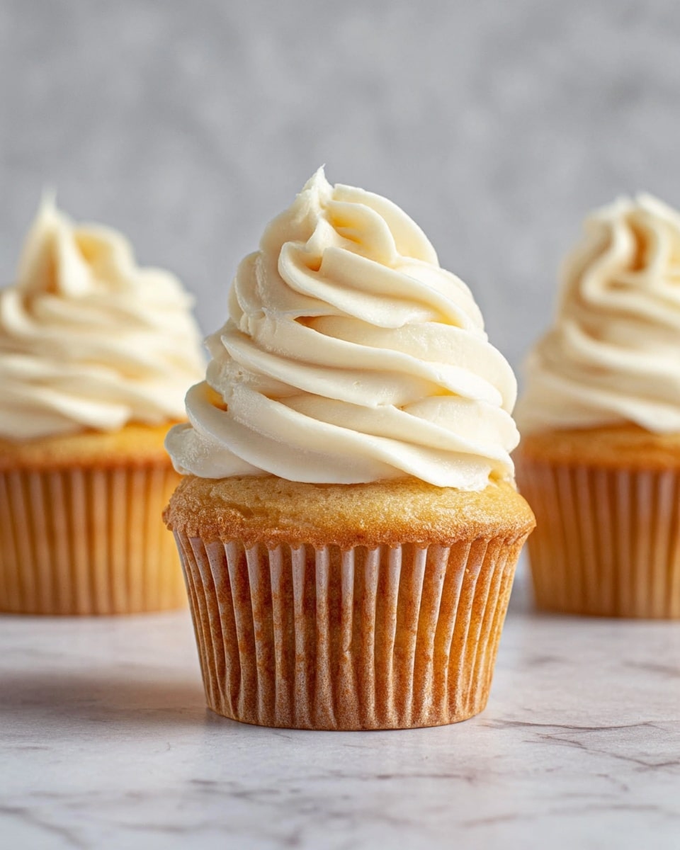 The image shows three cupcakes on a white marbled surface with a soft gray wall in the background. Each cupcake has a golden brown base with vertical lines from the baking paper. On top of each base, there is a thick, creamy white frosting swirled in a tall spiral shape with soft curves and a slightly glossy texture. The frosting is smooth and looks light and fluffy, standing out clearly against the golden brown base. The middle cupcake is the main focus, showing more detail in the texture of the frosting and cake. Photo taken with an iphone --ar 4:5 --v 7