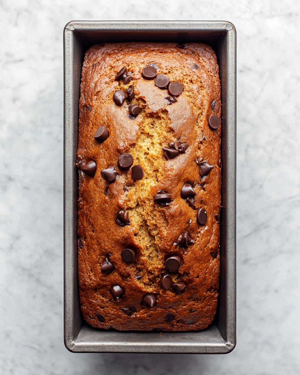This image shows a golden-brown loaf cake in a rectangular metal baking pan. The top is cracked slightly in the middle, revealing a soft inside filled with dark chocolate chips scattered all over the surface. The cake looks moist and fluffy with a rich brown color from the baking. The metal pan has sharp edges, and the whole scene is set on a white marbled background. photo taken with an iphone --ar 4:5 --v 7