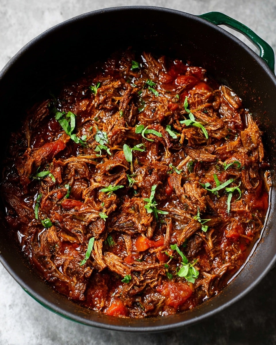 The image shows a close-up of shredded meat stew in a large black pot with green handles, sitting on a white marbled surface. The meat is dark brown and appears moist and tender, mixed with chunks of red tomato sauce and garnished with small bright green herb leaves scattered on top. The texture looks rich and saucy with the meat fibers clearly visible, and the sauce has a thick, slightly shiny appearance, coating the meat well. Photo taken with an iphone --ar 4:5 --v 7