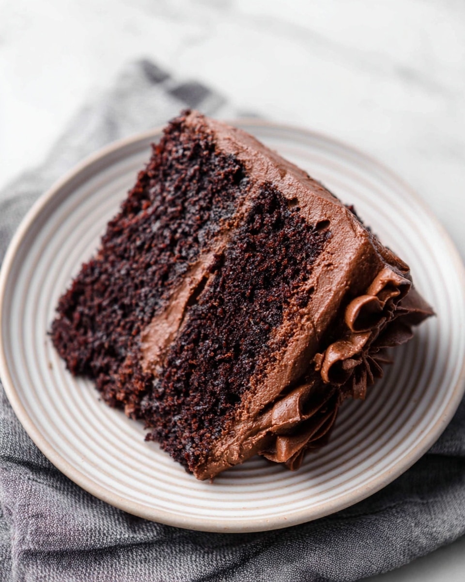 A slice of two-layer chocolate cake sits on a white plate with light circular ridges, placed on a white marbled surface. The cake layers are dark brown and moist with a rich texture. Between the two cake layers, there is a smooth layer of chocolate frosting. The same chocolate frosting covers the top and sides of the slice with swirled, slightly pointed peaks, showing a creamy texture. A gray striped cloth is slightly visible beneath the plate. photo taken with an iphone --ar 4:5 --v 7