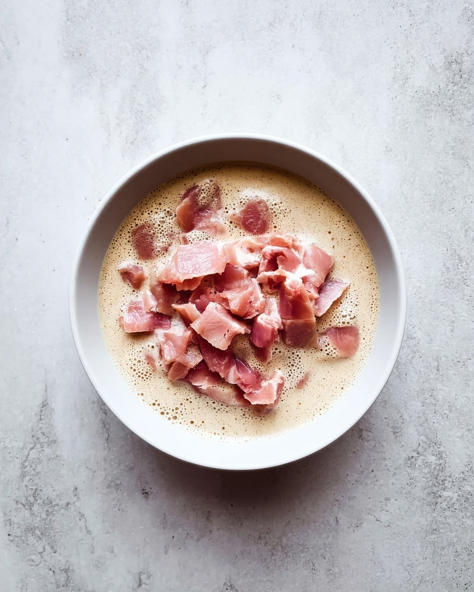 A white bowl sits on a white marbled surface, filled with a creamy beige liquid that has a frothy texture around the edges. On top of the liquid, there are many pieces of raw pinkish meat, layered unevenly in the center of the bowl. The contrast between the light creamy base and the pink meat pieces creates a simple yet raw look. photo taken with an iphone --ar 4:5 --v 7