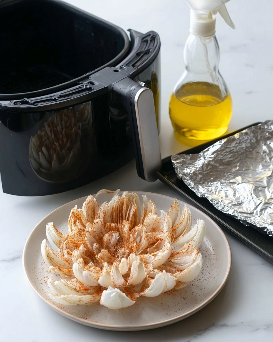 A plate with a large onion flower prepared for frying sits on a white marbled surface. The onion is cut into many thick layers that spread out like petals, each layer dusted with a light coating of flour or seasoning, giving a pale peach and white look with soft rough texture. Behind the plate, there is a black air fryer with the basket slightly open, showing its ridged, dark interior. Next to it, on the right side, is a clear spray bottle filled with yellow oil, resting on the surface. In front of these items, there is a dark rectangular tray with a sheet of crinkled aluminum foil laid across it. The background and surface are clean and white marbled for a bright, simple kitchen setup photo taken with an iphone --ar 4:5 --v 7