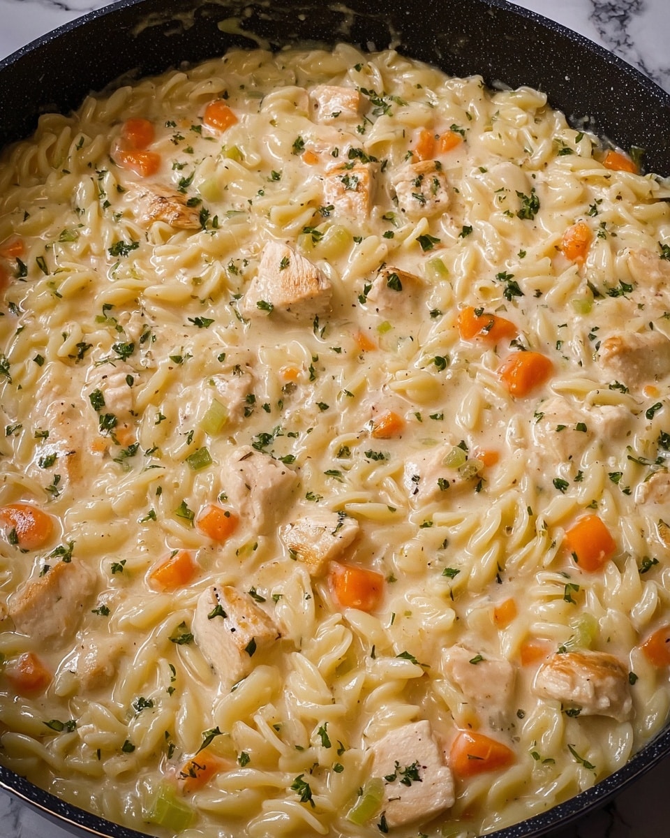 A close-up of a thick creamy dish in a dark pan showing one main layer filled with small pasta pieces, tender light beige chicken chunks, and diced orange carrots, along with bits of green celery. The creamy sauce is light yellow with a smooth texture, lightly sprinkled with green herbs on the surface. The background features a white marbled texture. photo taken with an iphone --ar 4:5 --v 7