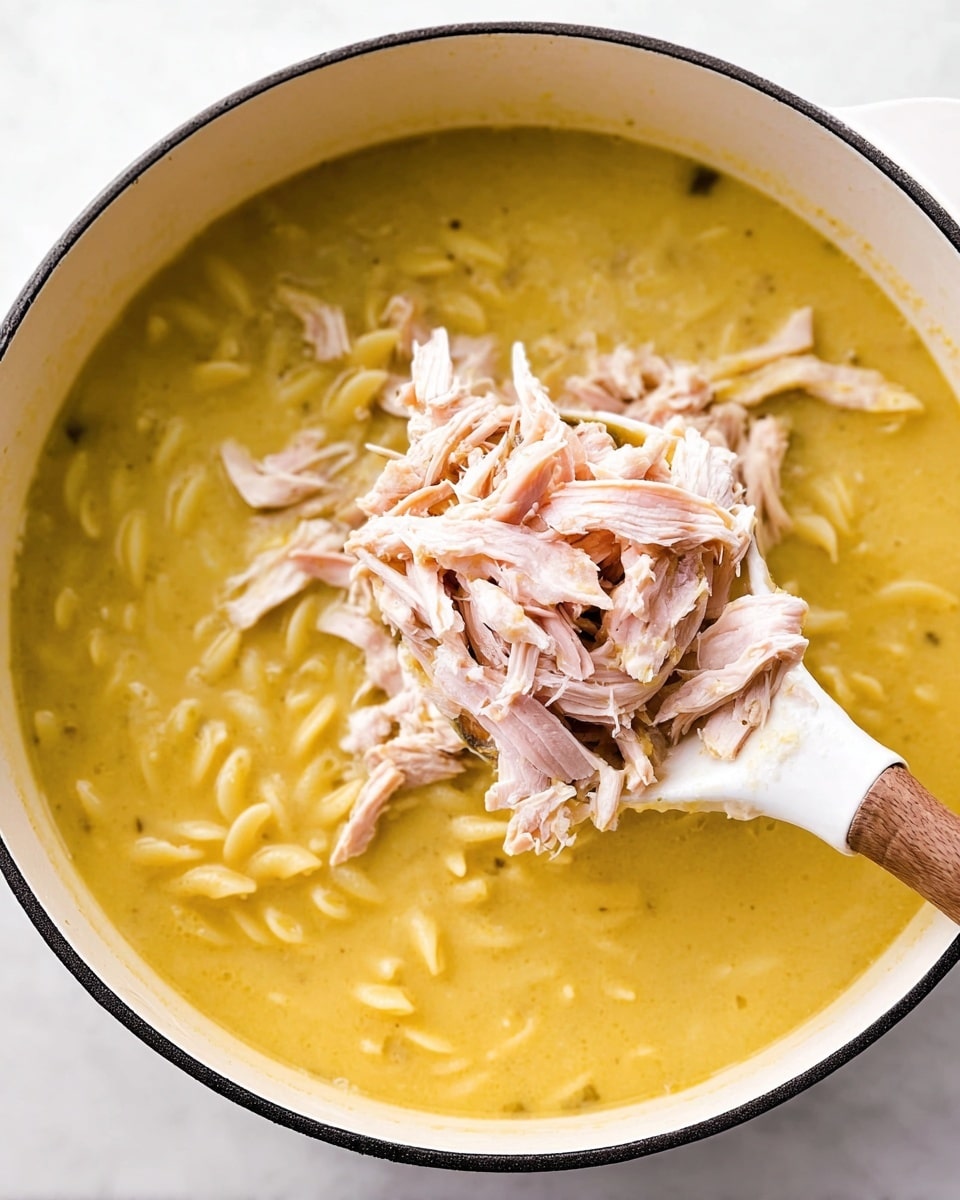 The image shows a white pot filled with yellow creamy soup that has some small pasta noodles mixed inside. On top of the soup, there is a heap of shredded light pink chicken pieces being held by a spoon with a wooden handle and a white scoop. The texture of the soup looks smooth and silky with the noodles adding slight thickness. The pot rests on a white marbled surface. photo taken with an iphone --ar 4:5 --v 7