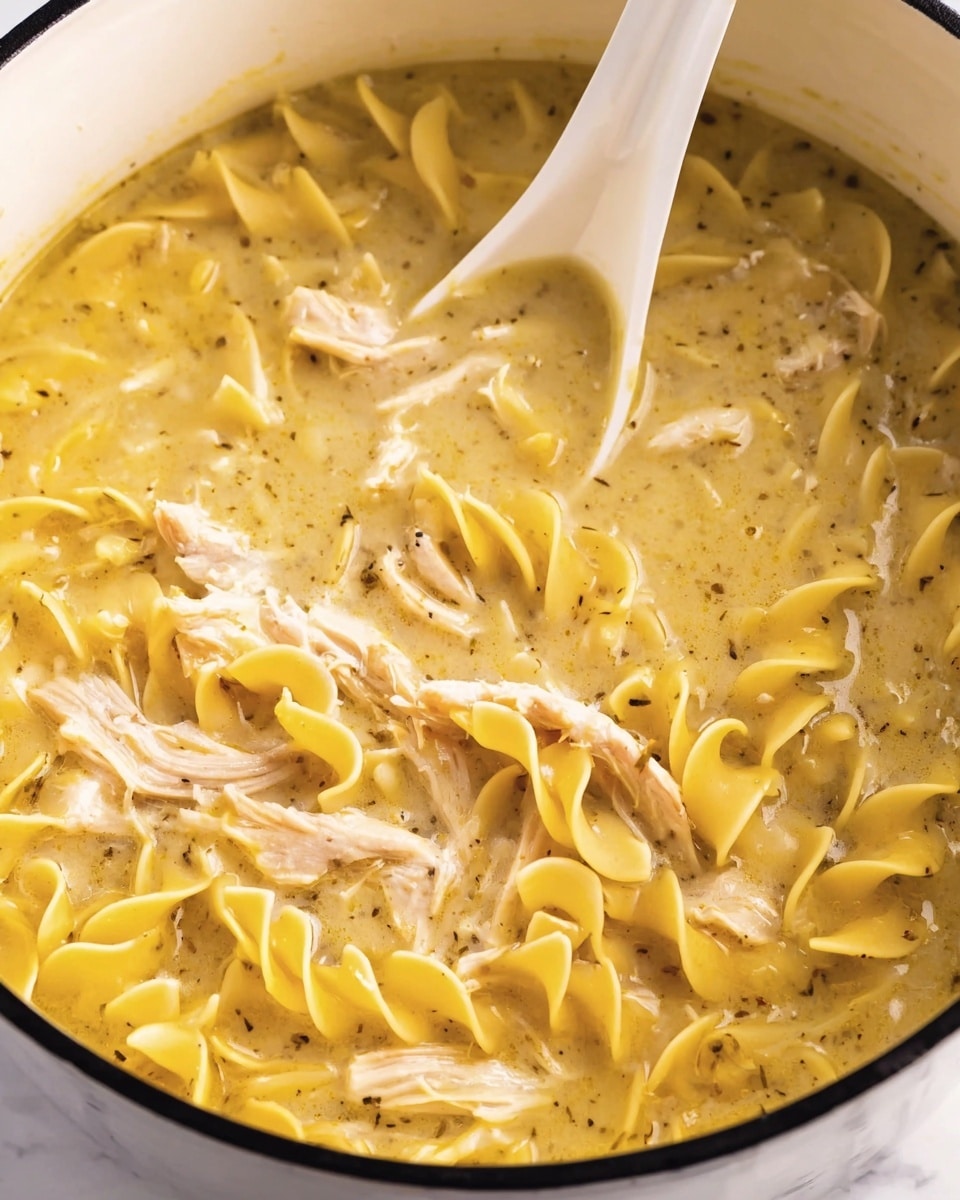 This image shows a close-up of a white pot filled with creamy yellow chicken noodle soup. The soup has three main layers: soft yellow egg noodles twisted through the broth, tender pieces of light brown shredded chicken scattered evenly, and a thick, smooth broth that looks rich and slightly speckled with black pepper. A white spoon is partially dipped into the soup, blending in with the creamy texture, with its handle rising from the top center of the pot. The background is a white marbled surface. photo taken with an iphone --ar 4:5 --v 7