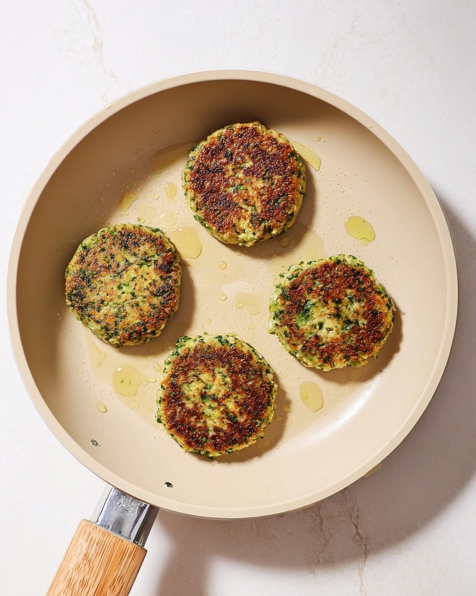 The image shows four round patties cooking in a beige frying pan with a wooden handle. The patties have a crisp, browned outside with green bits visible inside, suggesting herbs or vegetables mixed in. They are evenly spaced with small drops of cooking oil around them, creating a slight shine on the pan surface. The background is a white marbled texture. photo taken with an iphone --ar 4:5 --v 7