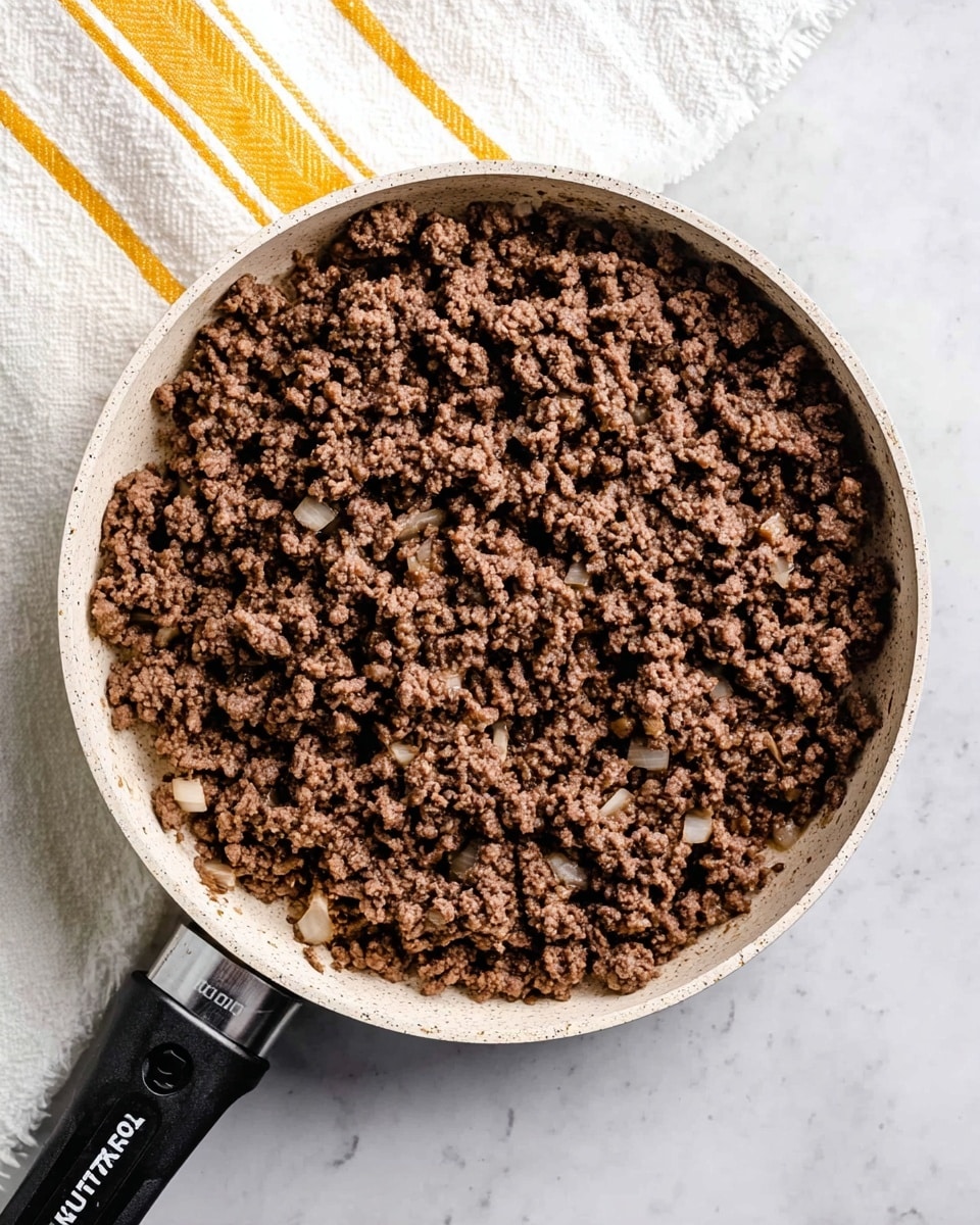 The image shows a white pan filled with cooked ground beef mixed evenly with small pieces of onion. The beef is brown and crumbly, covering the entire pan surface. The pan handle is black with white text on it. The pan is placed on a white marbled surface, and there is a white towel with yellow stripes partially visible near the top left of the pan. Photo taken with an iphone --ar 4:5 --v 7