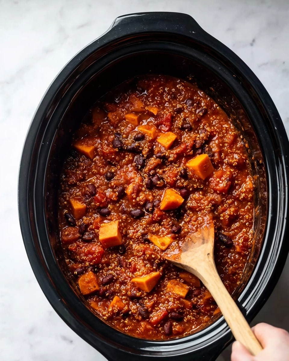 A black slow cooker filled with a thick reddish-brown stew made of small quinoa grains, black beans, diced orange sweet potatoes, and small chunks of tomato, all mixed in a rich sauce. A wooden spoon is inside the slow cooker, stirring the stew with a woman's hand holding the spoon. The background surface is white marbled texture. photo taken with an iphone --ar 4:5 --v 7