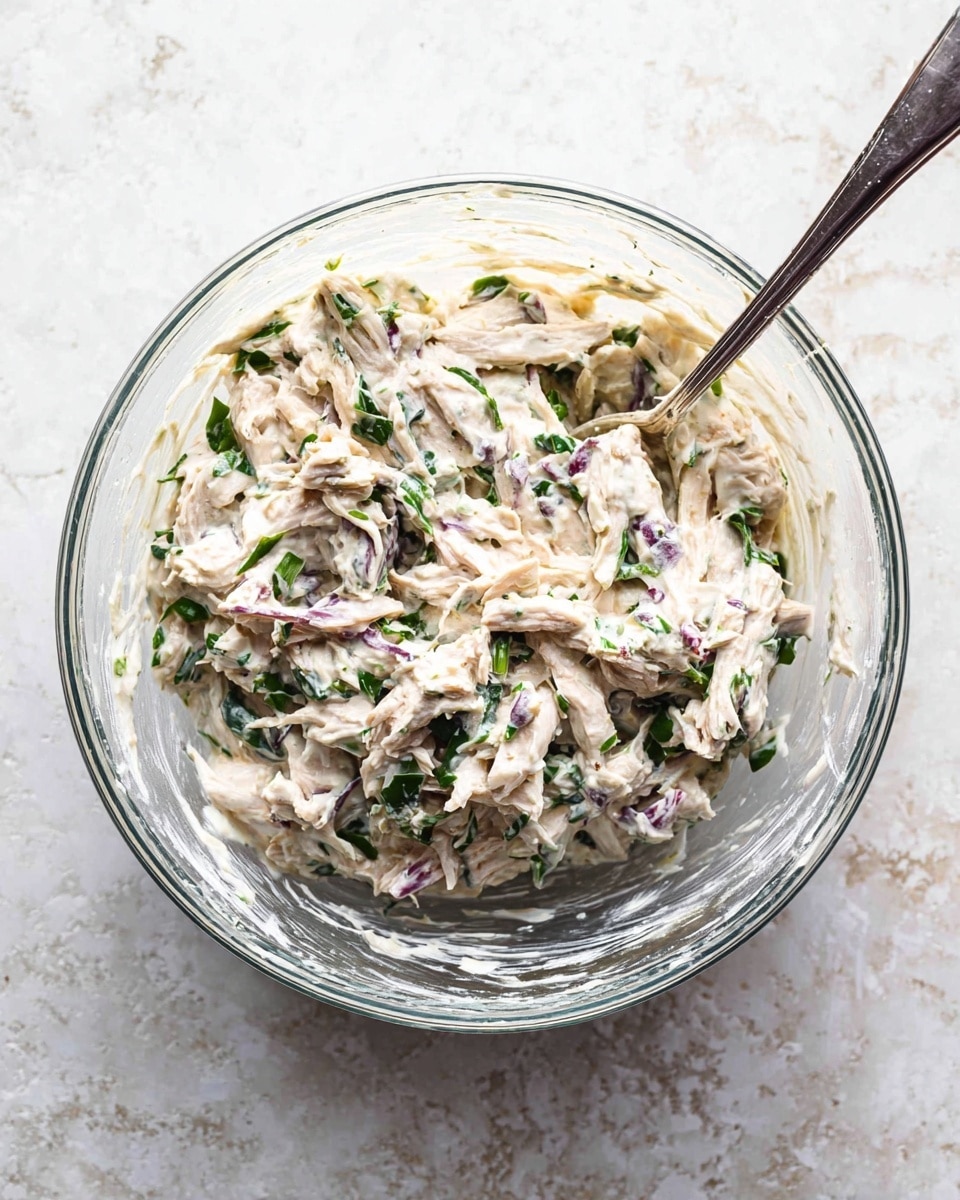 A clear glass bowl holds a creamy mixture with visible shredded white and light beige pieces that look like chicken, mixed with chopped green herbs and small purple bits, all surrounded by a white creamy sauce. A silver spoon is placed inside the bowl, resting at the side, and the bowl sits on a white marbled textured surface. The mixture has a thick, slightly chunky texture with green leaves and purple pieces scattered evenly throughout. photo taken with an iphone --ar 4:5 --v 7
