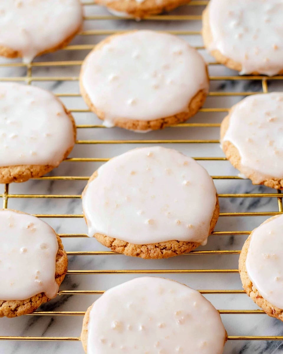 The image shows a collection of round cookies with a light brown base and a smooth, white glaze on top. The cookies are placed on a gold wire cooling rack that highlights their round shape and shiny iced surface. The white glaze covers each cookie completely, with slight drips forming at the edges. The cookies vary slightly in size, and the glaze has a glossy texture with some small air spots visible. The background features a white marbled texture that makes the cookies stand out clearly. photo taken with an iphone --ar 4:5 --v 7