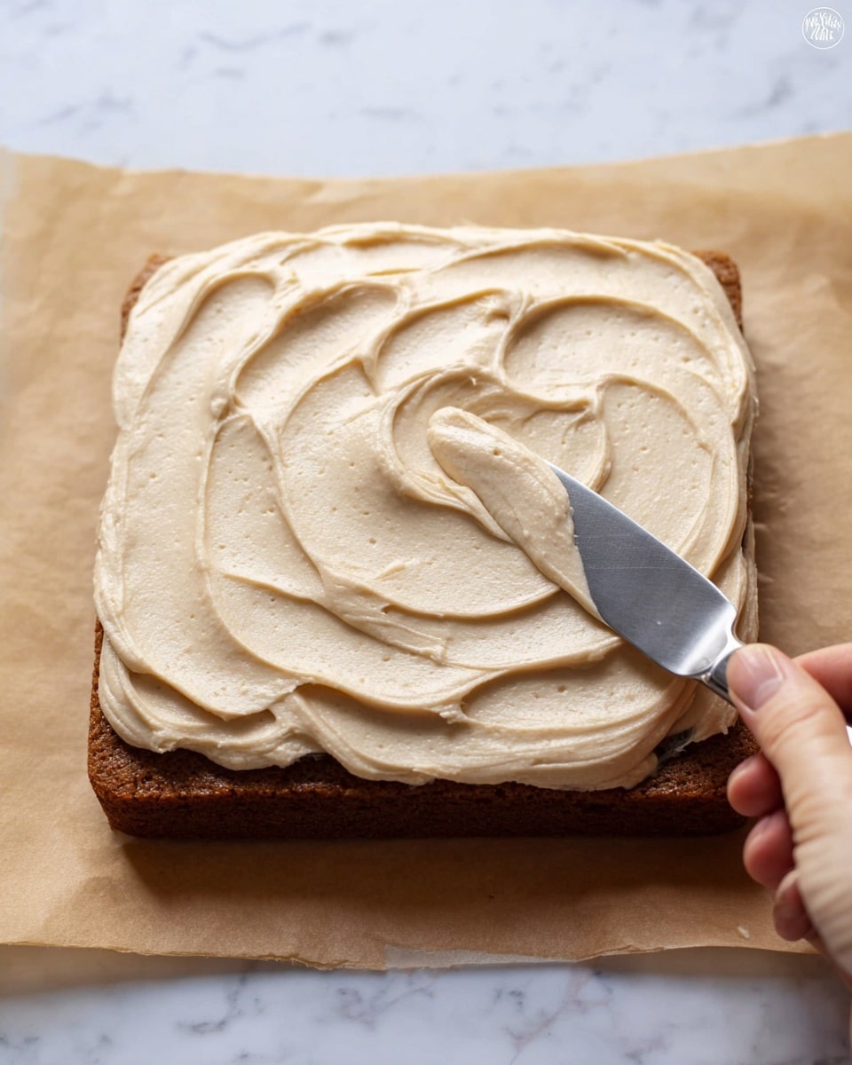 A close-up view of a single square layer of brown cake topped with a thick, creamy layer of light beige frosting being spread with a metal spatula held by a woman's hand. The frosting is textured with soft swirls and small air bubbles, covering the entire top surface of the cake, which sits on a piece of parchment paper over a white marbled surface. The photo taken with an iphone --ar 4:5 --v 7