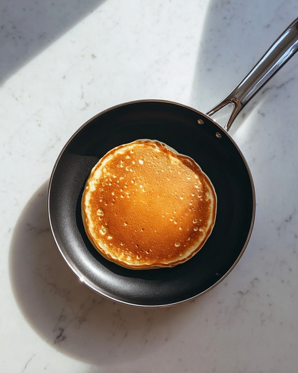 A single golden-brown pancake sits at the center of a black frying pan with a shiny, silver handle. The pancake has a smooth, round shape with tiny bubbles and a slightly lighter edge, showing it is cooked evenly. The frying pan rests on a white marbled surface, reflecting soft, natural light that falls in angular patterns. photo taken with an iphone --ar 4:5 --v 7