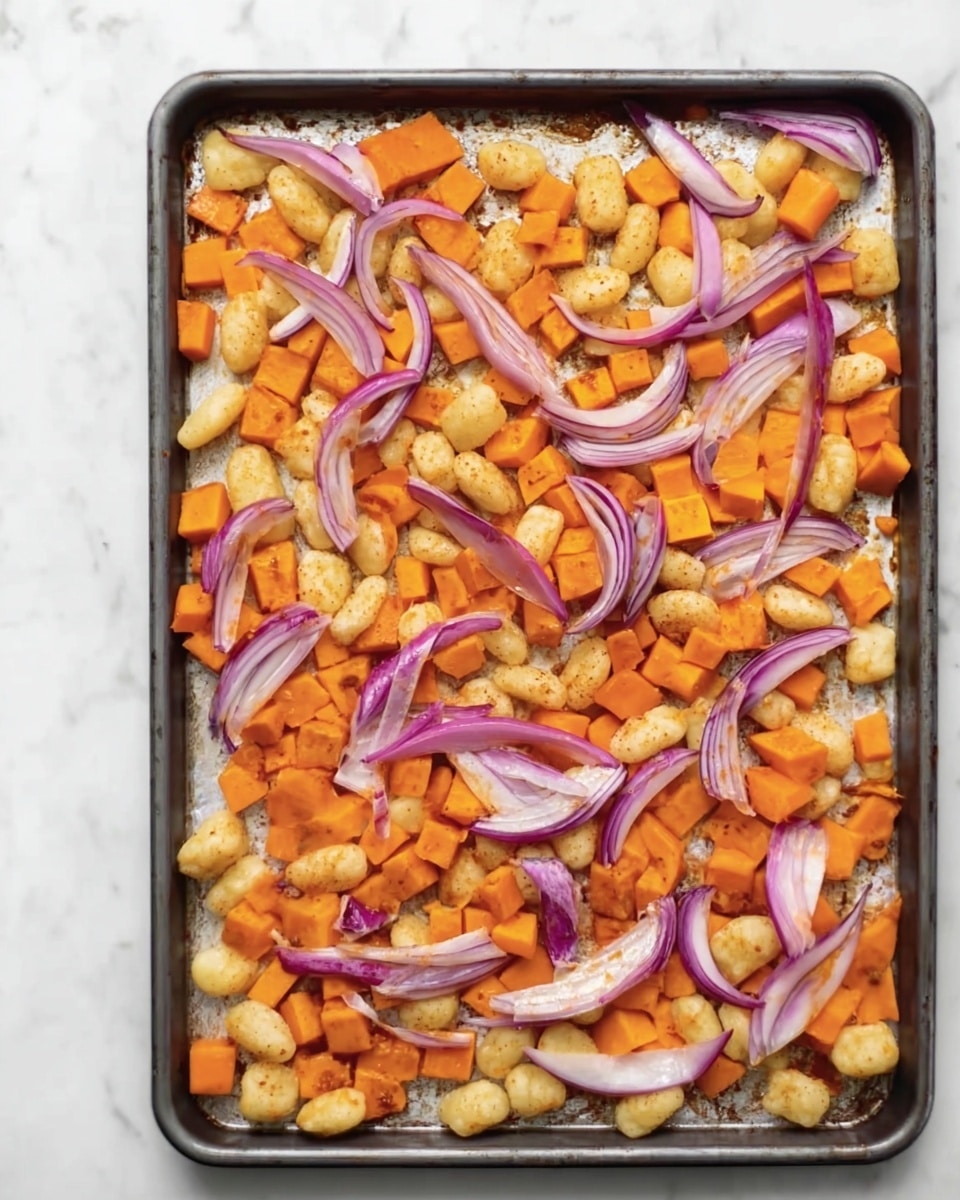 A baking tray filled with three main visible layers: the bottom layer is a slightly toasted white surface of the tray, the middle layer consists of many small golden-brown gnocchi pieces scattered evenly, and the top layer features bright orange sweet potato cubes and thin curved slices of purple-red onion spread over the gnocchi. The colors contrast well with each other, with the orange and purple standing out clearly against the pale gnocchi and tray. The scene is set on a white marbled surface. photo taken with an iphone --ar 4:5 --v 7