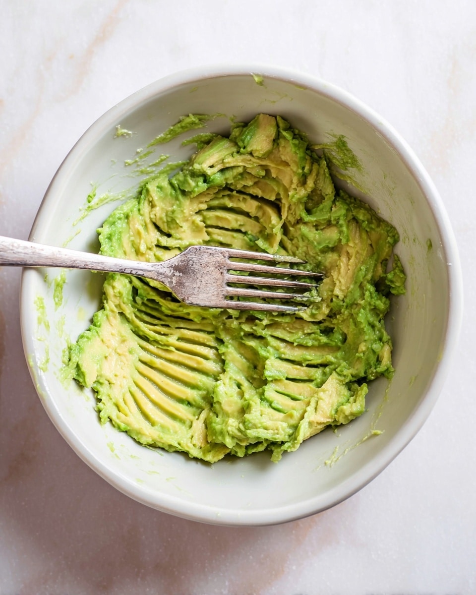 Inside a white bowl, there is mashed avocado being pressed with a silver fork. The avocado is a mix of light green and yellow shades with a soft, creamy texture. The bowl holds the avocado spread flat at the bottom, showing some ridges made by the fork. The white marbled surface can be seen below the bowl. Photo taken with an iphone --ar 4:5 --v 7