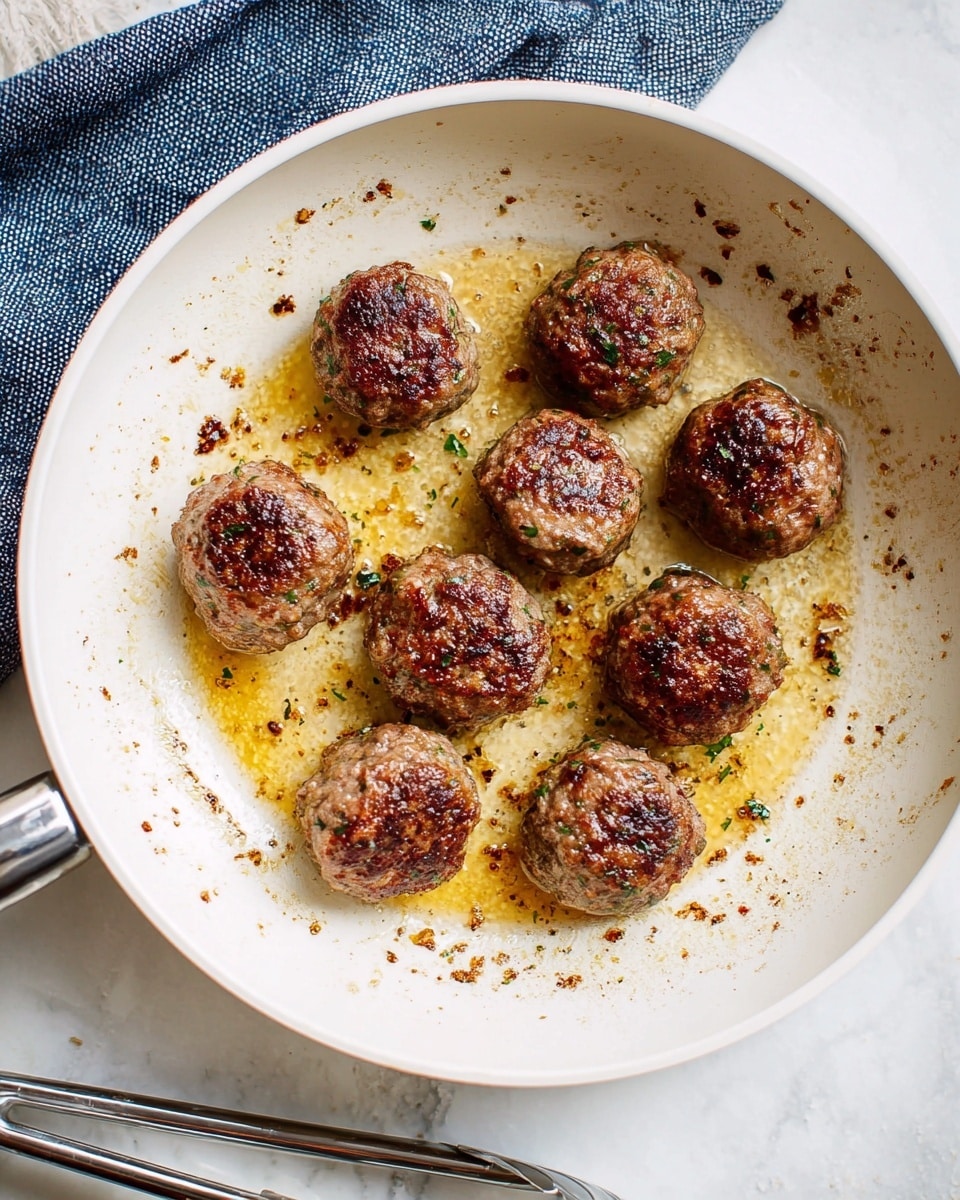 A white frying pan holds nine browned meatballs, arranged in a loose circular pattern with one in the center. The meatballs have a rich, textured surface with some green herbs visible, and they sit in a thin layer of golden brown cooking oil with small browned bits scattered around. The pan rests on a white marbled texture surface with a folded blue cloth visible on the left edge and shiny metal tongs placed below the pan. photo taken with an iphone --ar 4:5 --v 7