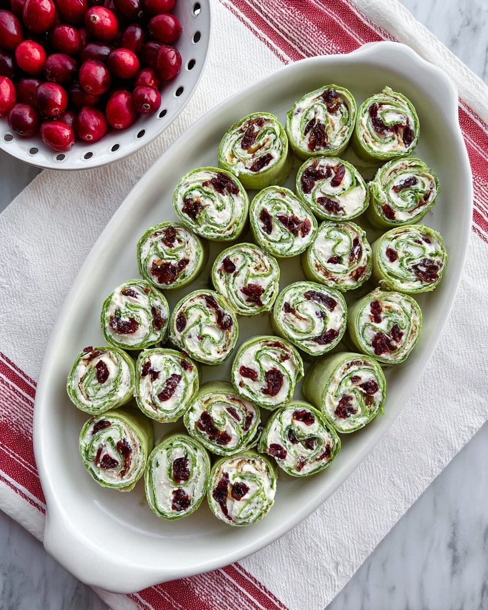 The image shows a white oval plate filled with 27 neatly arranged pinwheel-style rolls. Each roll has two visible layers: a green outer layer that looks soft like a spinach wrap, and an inner white layer of creamy filling spotted with dark red-brown bits, likely dried cranberries or similar fruit. The rolls are all uniform in size and tightly rolled. The plate sits on a white marbled surface, partially on top of a white towel with red patterned lines. A white colander with fresh red cranberries is visible in the upper left corner of the image. photo taken with an iphone --ar 4:5 --v 7