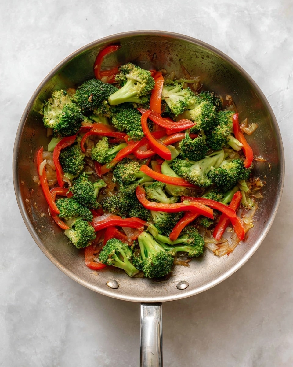 A close-up view of a metal frying pan with a shiny surface, containing a mix of cooked vegetables. The base layer in the pan shows cooked, slightly browned bits, while on top are bright green broccoli florets evenly spread across the pan. Interspersed with the broccoli are thin, long strips of red bell pepper, slightly glossy from cooking. The pan's handle extends downward and is metallic, reflecting light. The frying pan sits on a white marbled surface with faint grey veins, giving a clean kitchen look. Photo taken with an iphone --ar 4:5 --v 7