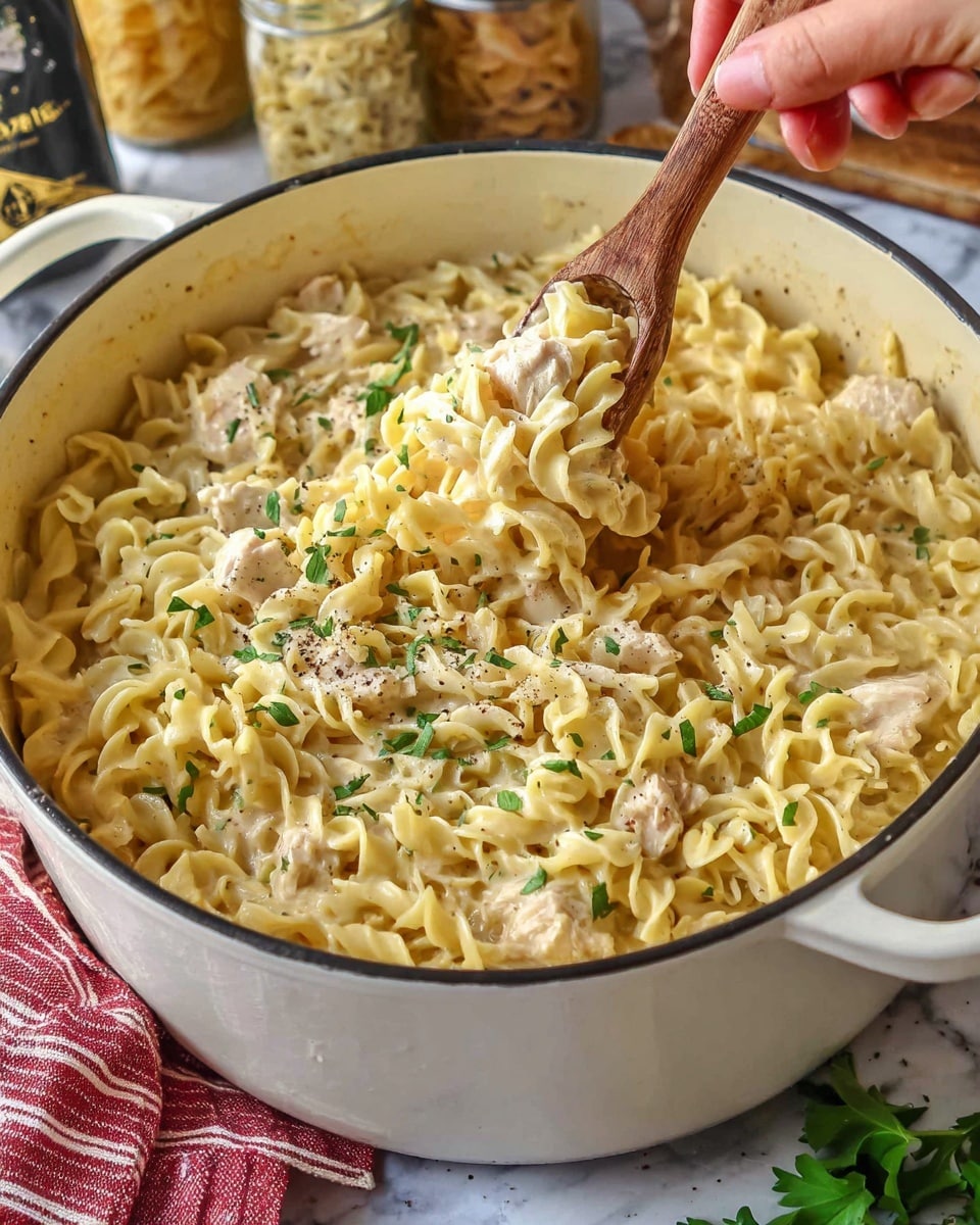 This image shows a large white pot filled with creamy cooked pasta with light yellow, curly noodles mixed with small pieces of light-colored chicken. The top layer has bits of green herbs and black pepper sprinkled evenly. A woman's hand is holding wooden tongs that lift some of the pasta and chicken from the pot. The pot is set on a white marbled surface with some green leaves and a folded red-striped cloth nearby. In the background, there are jars with pasta and ingredients slightly blurred. photo taken with an iphone --ar 4:5 --v 7