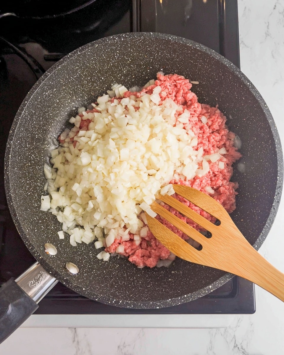 A top view of a speckled black cooking pan on a black stovetop showing two main layers inside the pan: the bottom layer is raw, pink ground meat spread flat, and the top layer is a large pile of small, white chopped onions covering much of the meat. A wooden spatula with slits rests on the right side of the pan, touching the onions and meat. The background is a white marbled texture. Photo taken with an iphone --ar 4:5 --v 7