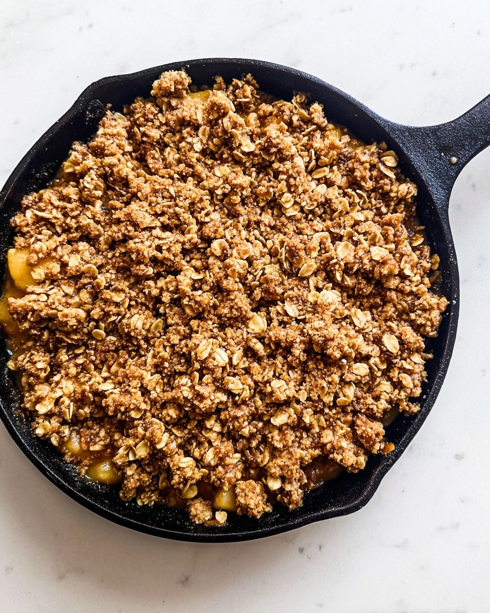 A round black skillet filled with a thick layer of golden brown oat crumble topping that looks crunchy and uneven in texture, showing small clusters of oats and crumb pieces scattered on top. Underneath, a hint of soft yellow fruit filling peeks through slightly at the edges. The skillet sits on a white marbled surface. photo taken with an iphone --ar 4:5 --v 7