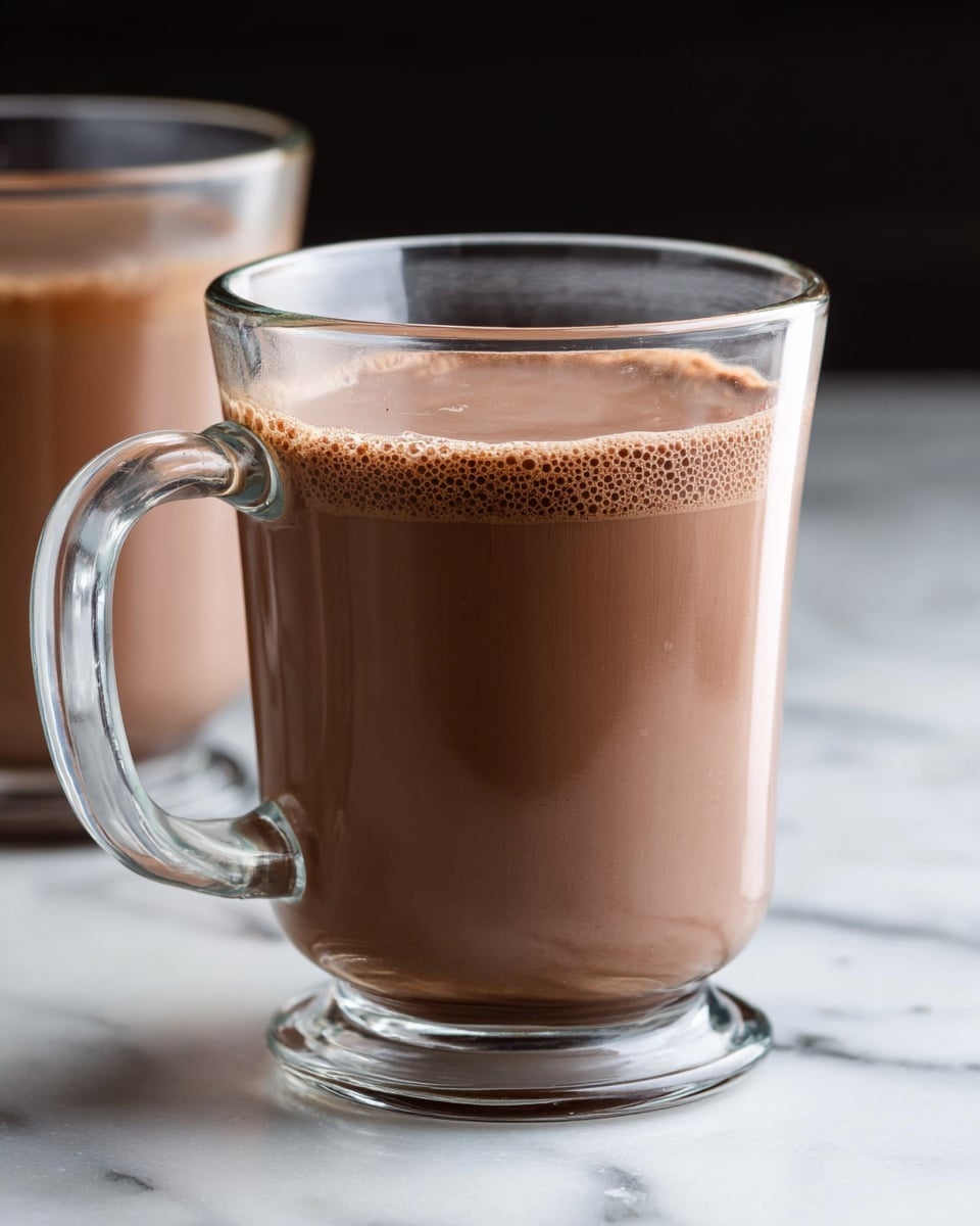 The image shows a clear glass mug filled with smooth, light brown hot chocolate, almost full to the top with a thin layer of tiny bubbles on the surface. The mug has a sturdy clear handle on the left side, and in the background, another similar glass mug is slightly out of focus. The mugs are placed on a white marbled surface with a black background behind them. photo taken with an iphone --ar 4:5 --v 7