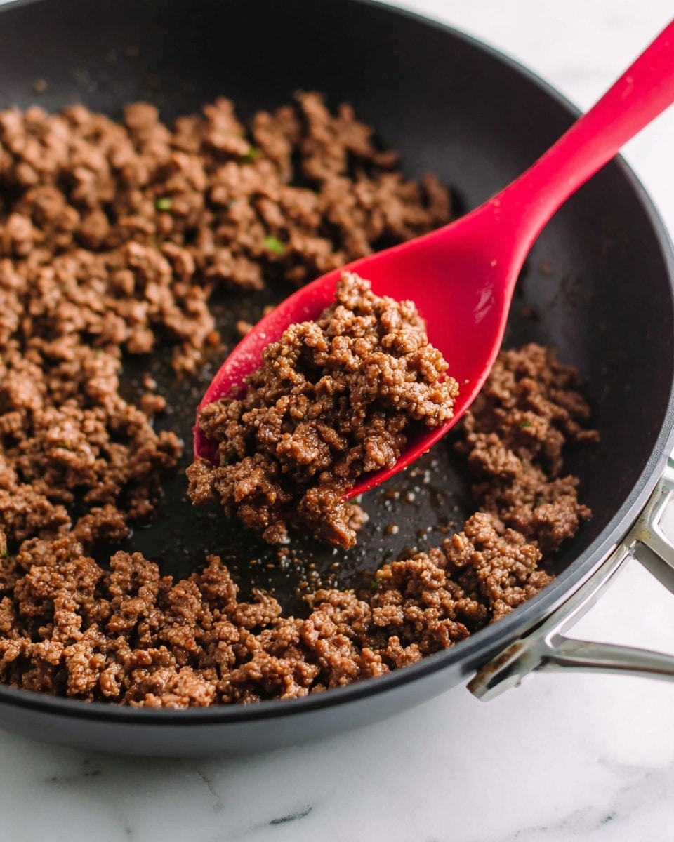 A close-up of a black skillet filled with cooked brown ground meat showing a slightly oily texture. A red spoon is scooping a portion of the meat from the skillet, with pieces of meat clinging to the spoon, highlighting its crumbly texture. The skillet rests on a white marbled surface, creating a clean and bright background. The metal handle of the skillet is visible in the upper right corner, contrasting with the dark pan and the vibrant red spoon. photo taken with an iphone --ar 4:5 --v 7
