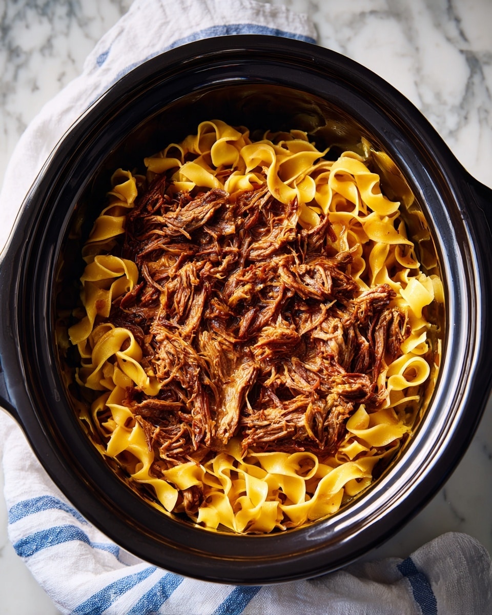Inside a black slow cooker, there is a mixture of two main layers: tender shredded brown meat and wide yellow pasta noodles. The pieces of meat are soft and stringy, tangled closely with the flat, broad noodles that have a smooth texture. The pasta and meat are evenly mixed together, showing a slight shine from sauce or cooking liquid. The pot sits on a white marbled surface, next to a white cloth with blue stripes. Photo taken with an iphone --ar 4:5 --v 7