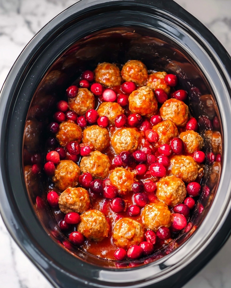 The image shows a black slow cooker filled with three layers: at the bottom, round meatballs coated with a shiny orange-red sauce, and scattered on top of the meatballs are bright red fresh cranberries, adding a pop of color and a smooth texture contrast. The slow cooker sits on a white marbled surface. Photo taken with an iphone --ar 4:5 --v 7