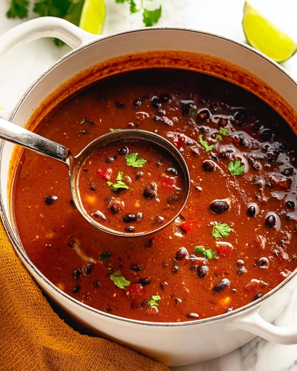 A white pot filled with dark reddish-brown soup with black beans, bits of red bell pepper, and small orange pieces, garnished with small green cilantro leaves on top. A shiny metal ladle dips into the thick, slightly shiny soup, showing a mix of smooth and chunky textures. The pot sits on a white marbled surface with a mustard-yellow cloth nearby and lime wedges in the background. The scene is bright and fresh with clear focus on the soup, photo taken with an iphone --ar 4:5 --v 7