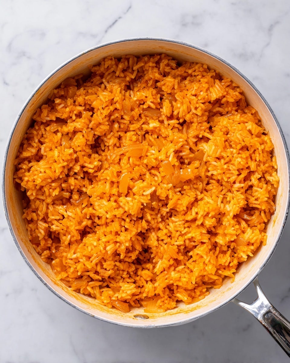 The image shows a close-up of a white pot filled with cooked rice that is bright orange in color. The rice appears fluffy with some small bits of cooked onions mixed in. The orange color is spread evenly through the rice, giving the dish a rich and warm look. The pot is placed on a white marbled surface, and the edge of the pot has a silver rim with a simple handle on the right side. There are no other visible ingredients or garnishes in the pot. Photo taken with an iphone --ar 4:5 --v 7