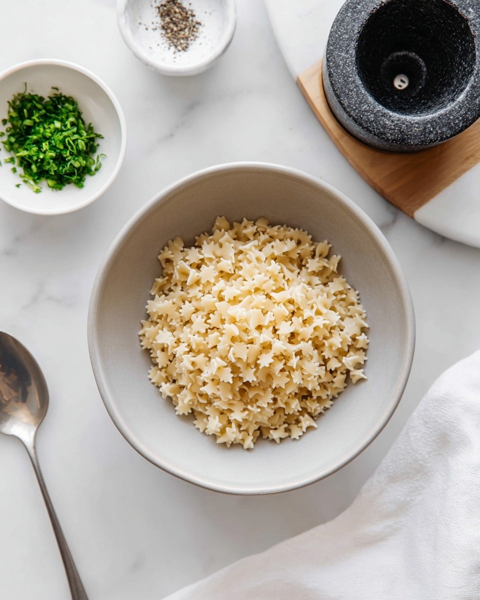 A simple bowl of cooked pasta shaped like small flowers and ridged spirals, placed in a light gray bowl with a smooth texture, sitting on a white marbled surface. To the top left, there is a small white bowl filled with chopped green herbs, and nearby a white spice container partially in view. On the right side, a black stone pepper grinder is placed on a white wooden board. A silver spoon lies to the left of the bowl, and a white cloth napkin is partly visible on the right side of the frame. The image has soft natural lighting and a clean, minimal look. photo taken with an iphone --ar 4:5 --v 7