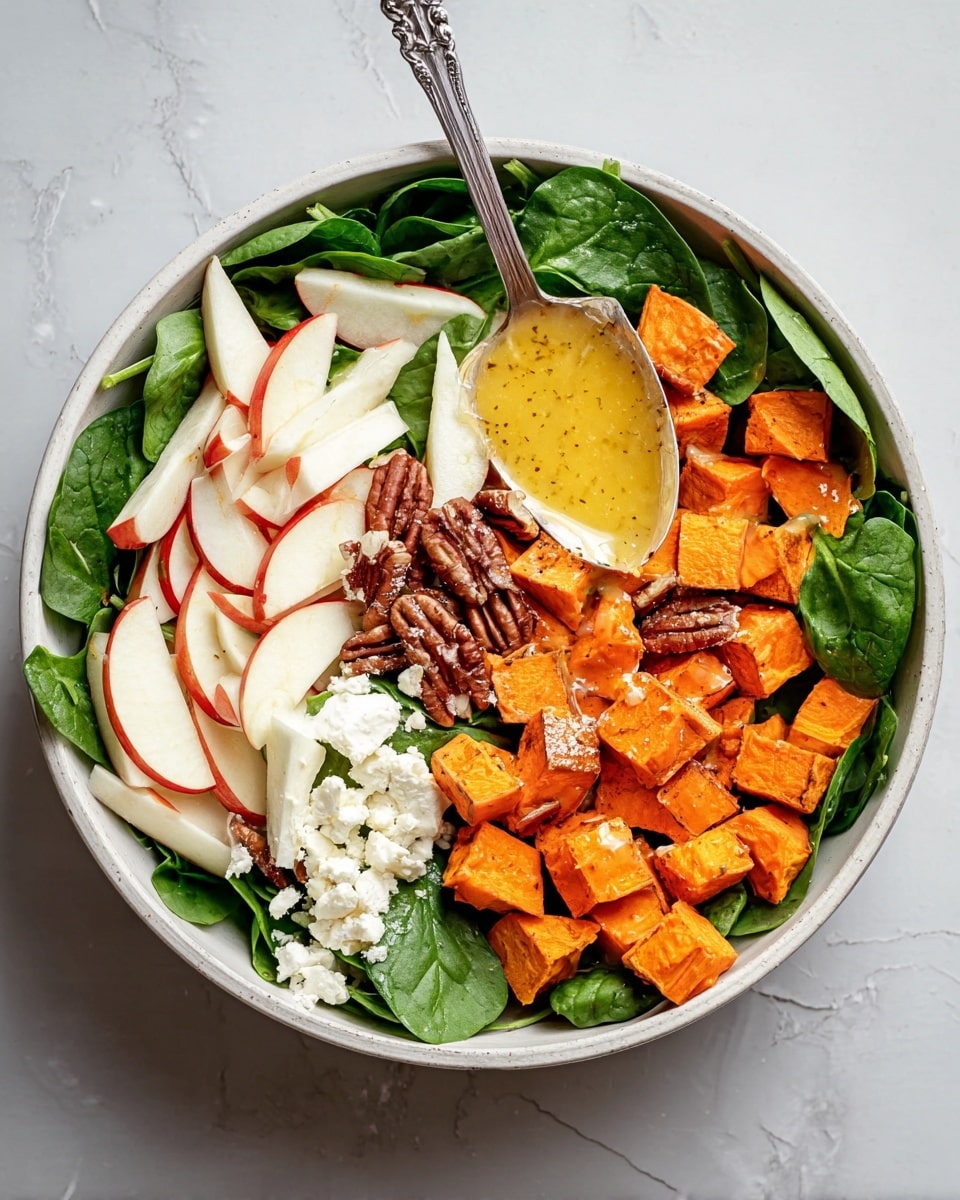 The dish is a warm salad served in a round white bowl on a white marbled surface. The bottom layer is fresh dark green spinach leaves spread evenly. On top of the spinach are medium-sized roasted orange sweet potato cubes placed mainly on one side. Next to the sweet potatoes is a pile of white, thinly sliced apple sticks with red skin edges. In between and slightly scattered are brown roasted pecans. There is also a small mound of crumbly white cheese near the spinach. A shiny silver spoon with a yellow, slightly chunky dressing is positioned over the bowl, ready to drizzle sauce over the salad. Photo taken with an iphone --ar 4:5 --v 7