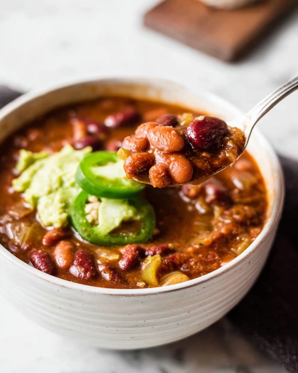 A white textured bowl filled with a thick, brown chili soup containing red and light brown beans, finely chopped onions, and small pieces of tomato. On the surface, there are smooth dollops of light green avocado chunks and bright green jalapeño slices. A spoon holds a scoop of the chili, showing the mix of beans, onions, tomatoes, and a dollop of avocado. In the background, wooden textures and a white marbled surface are visible. photo taken with an iphone --ar 4:5 --v 7