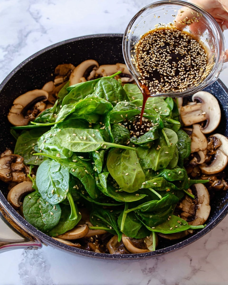 A black pan sits on a white marbled surface filled with two layers: the bottom layer has cooked brown and cream mushroom slices spread out evenly, while the top layer has fresh bright green spinach leaves piled in the center. A dark sauce with sesame seeds is being poured over the spinach from a clear glass bowl held by a woman's hand above the pan. The scene has a fresh and lively feel. photo taken with an iphone --ar 4:5 --v 7
