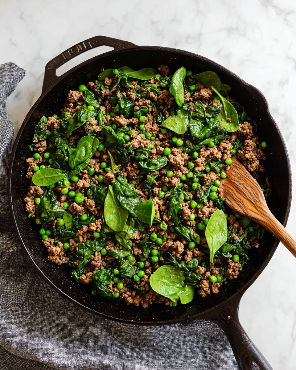 A black cast iron skillet filled with cooked ground meat mixed with scattered bright green peas and fresh spinach leaves on top, showing a combination of small brown textured bits and vibrant green layers, with a wooden spatula partially inserted under the food at the right side, all placed on a white marbled surface with a grey cloth nearby. Photo taken with an iphone --ar 4:5 --v 7
