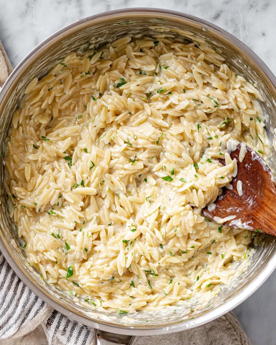 A close-up view of a large silver pot filled with creamy orzo pasta mixed with a white sauce, small bits of green herbs scattered throughout, and a wooden spoon partially submerged in the mixture on the right side. The orzo grains are plump and coated evenly with the sauce, creating a thick, rich texture. The pot rests on a white marbled surface with a striped cloth partially visible beneath it. Photo taken with an iphone --ar 4:5 --v 7