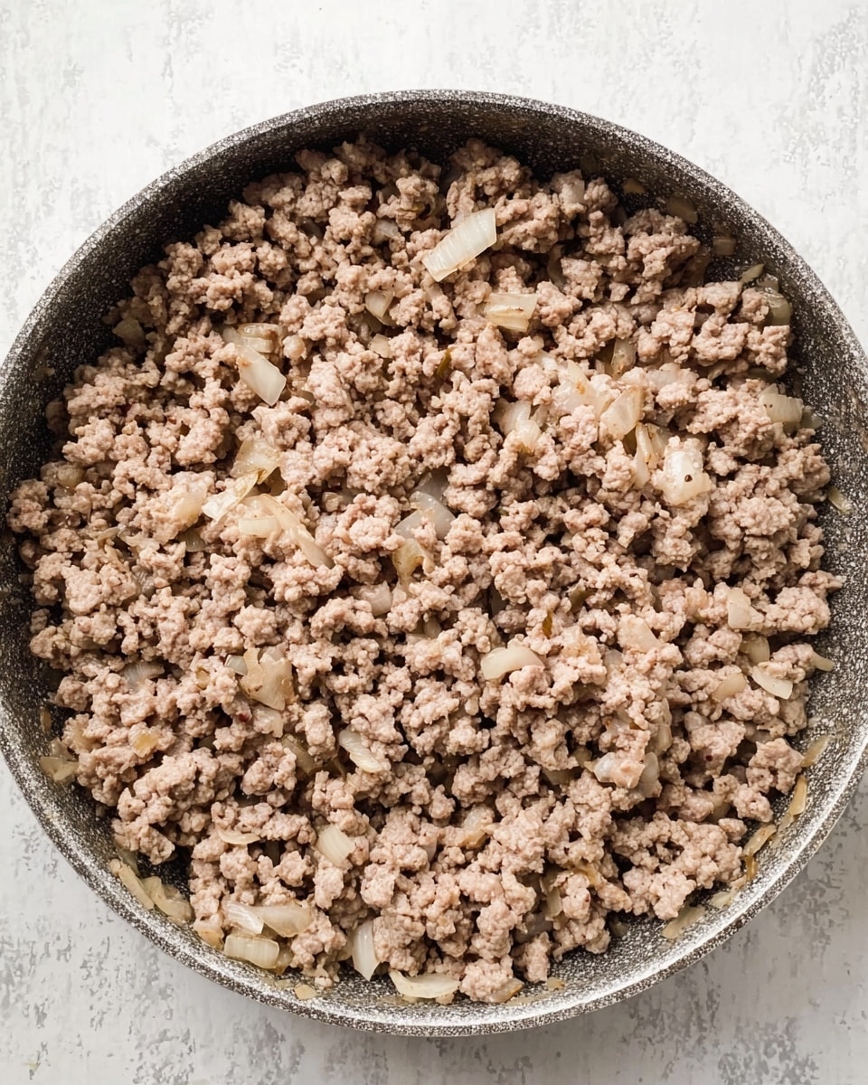 The image shows one layer of cooked ground meat mixed with small pieces of sautéed onions in a large shallow pan. The ground meat is light brown and crumbly, filling the pan evenly, while the onions are small, translucent, and scattered throughout the meat. The pan has a textured surface and occupies the entire frame. The background is a white marbled texture. Photo taken with an iphone --ar 4:5 --v 7