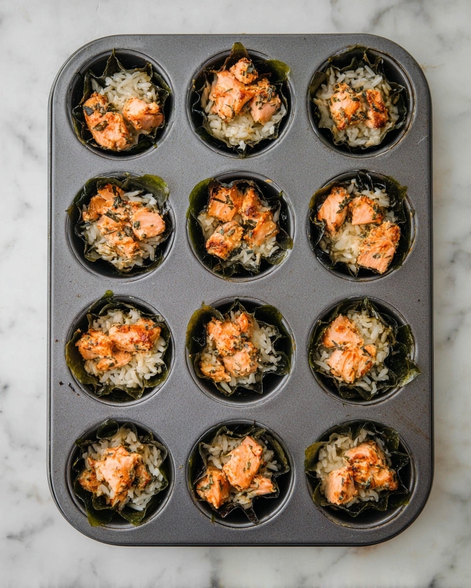 A dark-colored muffin tray holds twelve round sections, each lined with a dark green seaweed sheet forming a loose cup. Inside each seaweed cup is a base layer of white rice with a slightly sticky texture. On top of the rice, there are small chunks of cooked salmon, which have a pinkish-orange color with some browned edges, giving a cooked, tender look. The muffin tray sits on a white marbled surface, highlighting the contrast between the metal tray and the food inside. photo taken with an iphone --ar 4:5 --v 7