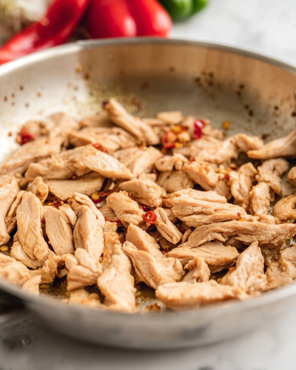 The image shows a close-up of a silver pan filled with small strips of cooked light brown meat, slightly shiny with a few darker spots of seasoning or chili flakes. The meat pieces are spread evenly across the pan's surface, showing a tender texture. Behind the pan, some blurred vegetables, including a bright red bell pepper, add subtle color contrast. The pan is placed on a white marbled surface. Photo taken with an iphone --ar 4:5 --v 7
