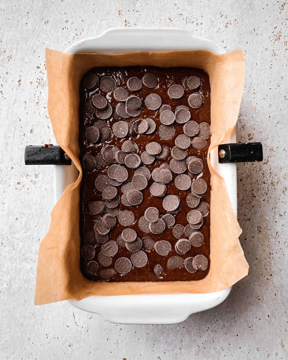 A white rectangular baking dish lined with parchment paper holds a single thick layer of dark brown chocolate batter sprinkled with many small round chocolate discs scattered unevenly on the surface. The dish rests on a white marbled textured surface, and two black clips are attached on the longer sides of the parchment paper, securing it in place. The batter looks smooth and shiny with a slightly uneven top, ready to be baked. photo taken with an iphone --ar 4:5 --v 7