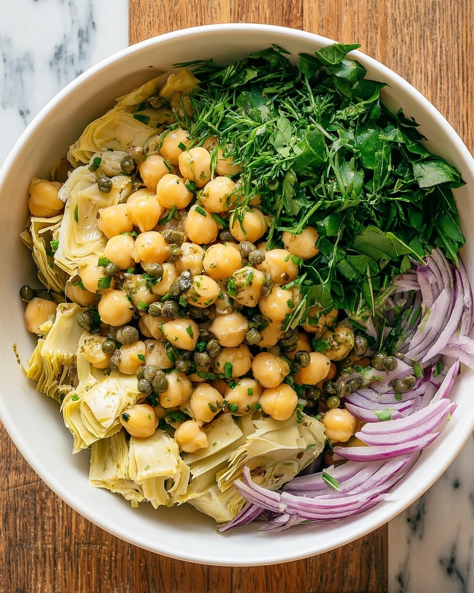 A white bowl filled with a chickpea salad showing three main layers: the bottom layer has thin slices of purple onion scattered around the bowl, the middle layer is a generous serving of light yellow chickpeas mixed with artichoke hearts that have soft, pale yellow leaves, and the top layer is made up of fresh green herbs including chopped chives, torn basil leaves, and capers mixed with a light green oily dressing that glistens under the light, all placed on a wooden surface with a white marbled texture background photo taken with an iphone --ar 4:5 --v 7