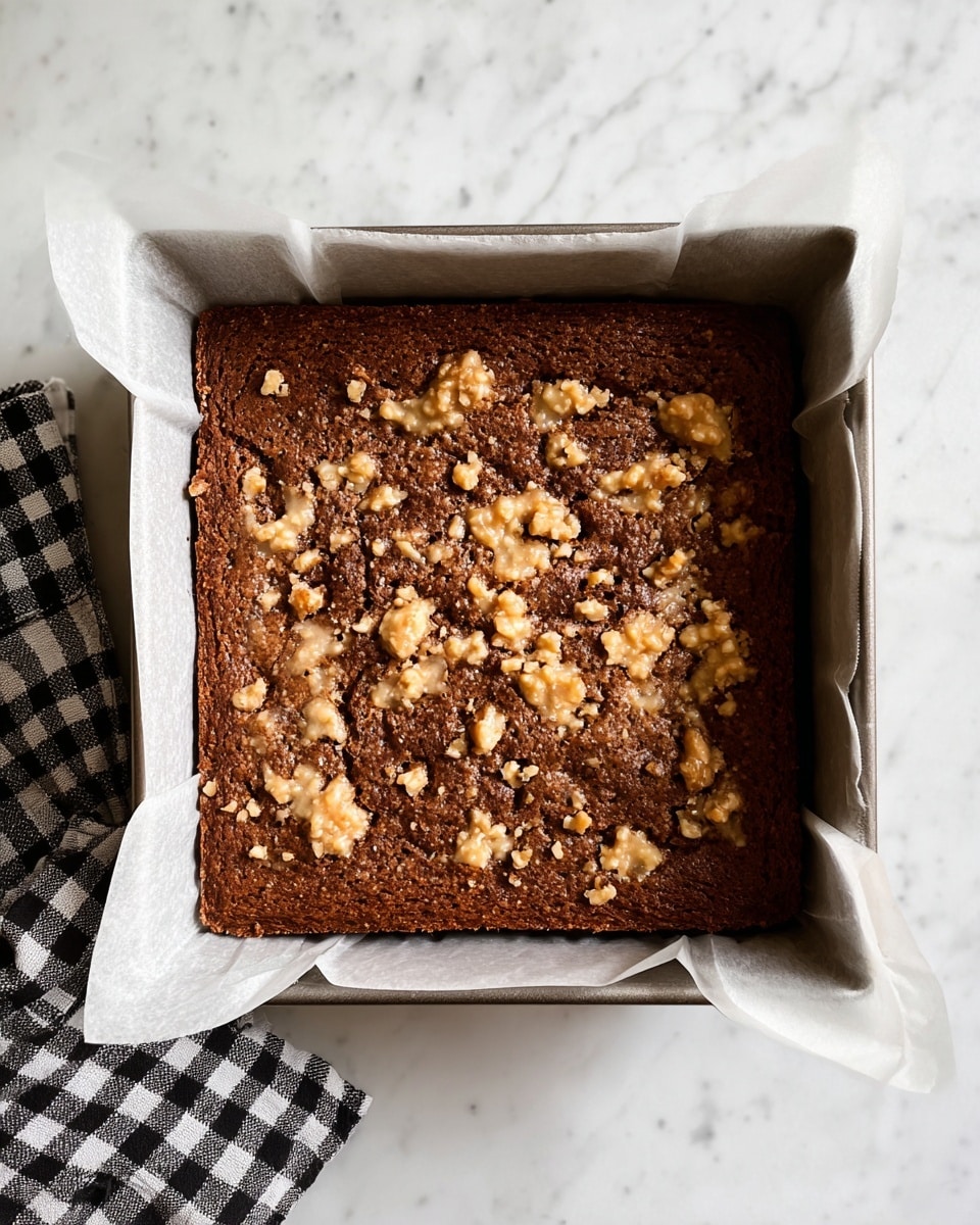 A square baked cake with a rich brown color forms the base layer, topped with uneven patches of a light beige mixture that has a slightly rough and chunky texture scattered across the surface. The cake is neatly placed inside a baking tray lined with white parchment paper that folds over the edges. The tray rests on a white marbled surface with a black-and-white checkered cloth partially visible beneath the bottom left corner of the tray. Photo taken with an iphone --ar 4:5 --v 7