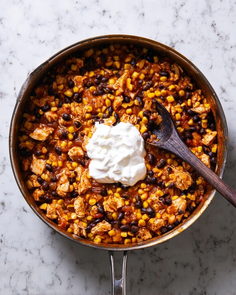 A metal pan filled with a colorful mixture of yellow corn, black beans, and small pieces of light orange cooked chicken, all combined in a reddish sauce. In the center of the pan, there is a large dollop of white sour cream sitting on top of the mixture. A wooden spoon with a dark handle is inside the pan on the right side, touching the food. The pan is placed on a white marbled surface. photo taken with an iphone --ar 4:5 --v 7